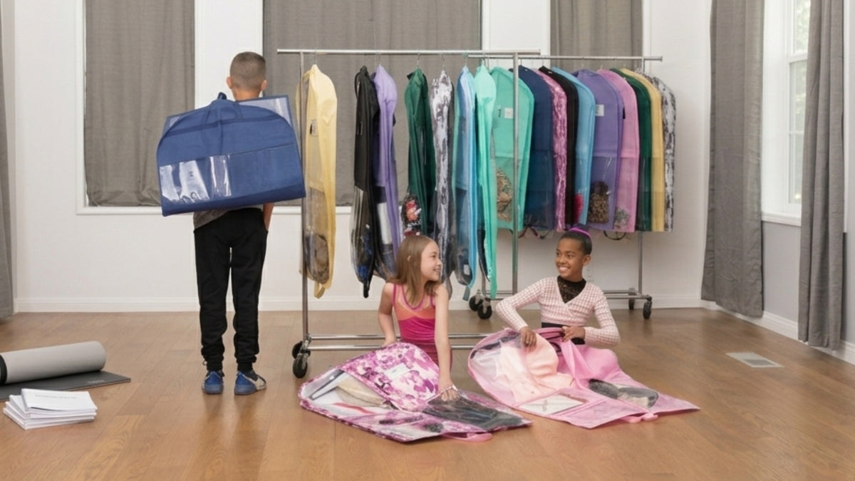 A rack of dance costume garment bags with clear panels in a studio, with two young dancers packing costumes into garment bags on the floor